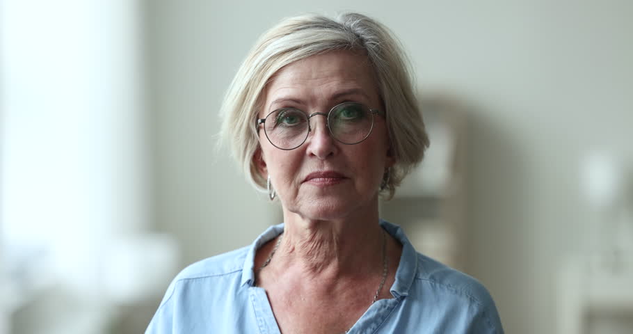 Serious pretty blonde senior woman wearing stylish round glasses, looking at camera, getting happy, cheerful, smiling with perfect white teeth. Elderly retired beauty care model head shot portrait Royalty-Free Stock Footage #1104029193