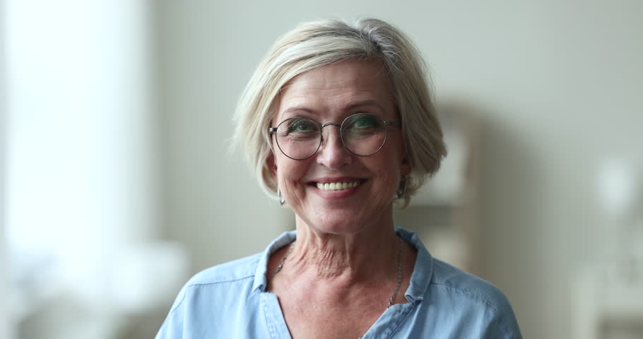 Serious pretty blonde senior woman wearing stylish round glasses, looking at camera, getting happy, cheerful, smiling with perfect white teeth. Elderly retired beauty care model head shot portrait