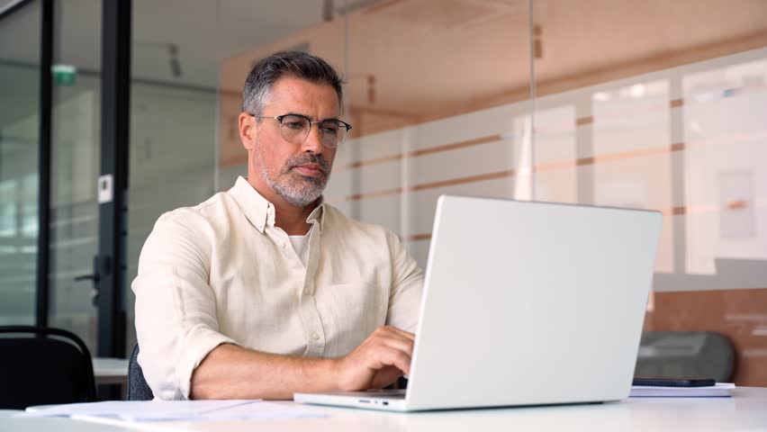 Mature Indian or Latin business man ceo trader using computer, typing, working in modern office, doing online data market analysis, thinking planning tech strategy looking at laptop with copy space. Royalty-Free Stock Footage #1104036179
