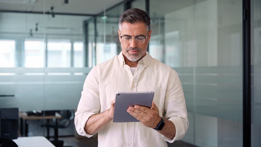 Happy Latin Hispanic bearded stylish mature adult professional business man, smiling Indian senior businessman CEO holding digital tablet using fintech tab application standing inside company office. Royalty-Free Stock Footage #1104036187