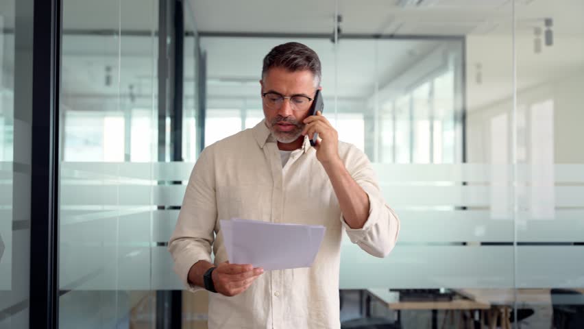 Mature Hispanic man talking by mobile cellphone at work in modern office. Middle aged Latin or Indian businessman walking, having call on smartphone with business partners and looking at documents. Royalty-Free Stock Footage #1104036193