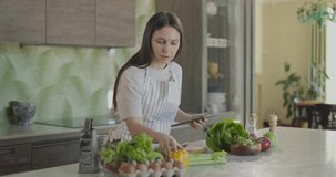 Girl in apron in kitchen is looking for recipe for healthy dish on tablet. Girl does not find recipe and angry. Slow motion