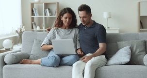 Hispanic couple spend free time seated on cozy couch using laptop, enjoy leisure on internet, buy goods, discuss e-services purchase, planning vacation, buy tickets, booking hotel. E-commerce clients