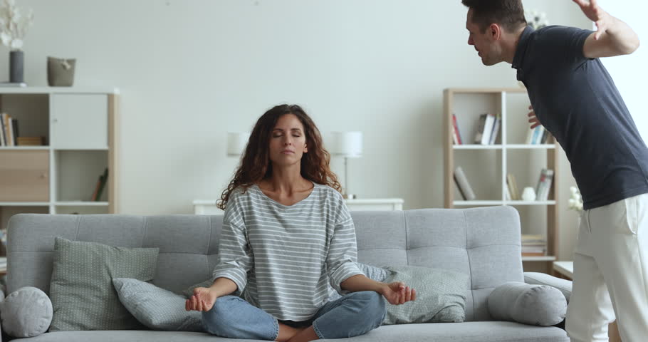 Boyfriend screams at girlfriend while she sits cross-legged do yoga exercises, deep in meditation practice, relieving stress, ignoring hysterics of young husband. Bad relationships, quarrel, jealousy Royalty-Free Stock Footage #1104040045
