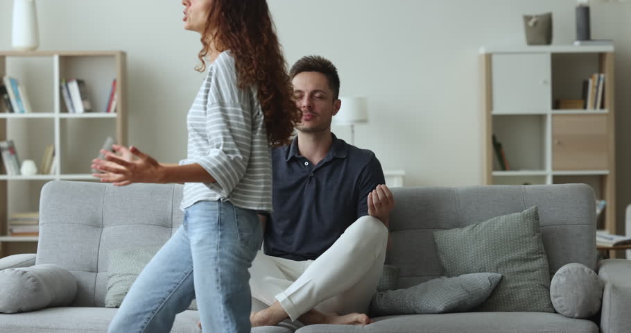 Husband sit cross-legged on couch doing meditation, express patience, not listening screams and hysterics of his wife. Spoiled relations, lack of harmony and understanding between spouses, jealousy Royalty-Free Stock Footage #1104040063