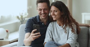 Happy Hispanic young couple spend weekend at home sit on couch at living room with cell phone, having fun with new mobile application, laughing on funny on-line videos in social media, chuckle at joke