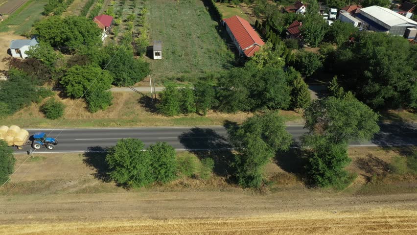 Above view, tractor as dragging two trailers piled up with round bales of straw going over asphalt road, path between cultivated fields and rural countryside.