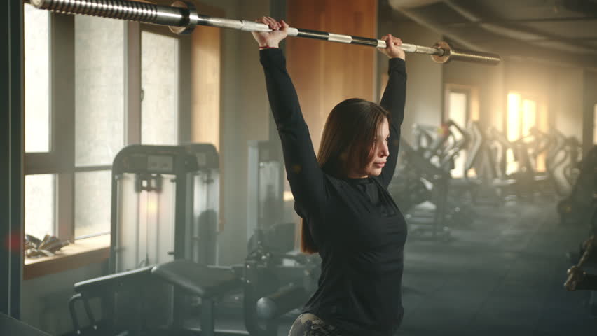 Medium shot of young cute smiling woman performing overhead press with Olympic barbell in gym. Woman lifts barbell overhead and holds it several seconds. High quality 4k footage Royalty-Free Stock Footage #1104044793