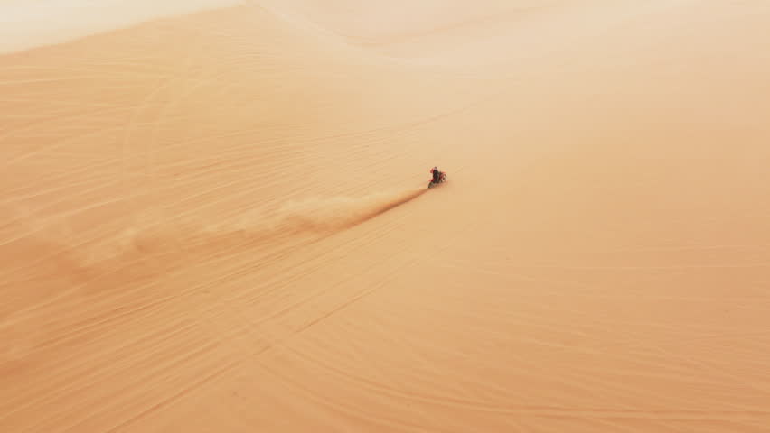 Skilled motocross rider drives on sand dune 4K. Dirt biker off roading on sand dunes in desert. Aerial view drone shot of extreme sport with man riding fast by sand dunes with scenic sand dust behind