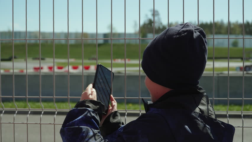 Boy is trying to shoot video on smartphone of car passing through gaps in metal fence along race track. On cool spring day, child uses their wits to film blog post.