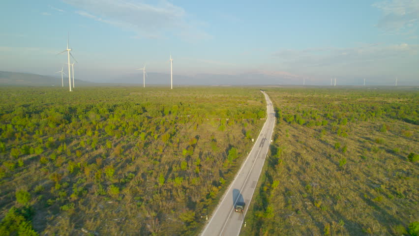 AERIAL: SUV car travelling on asphalt road and passing by working wind turbines. Scenic view of wind farm with scattered rotating windmills and road leading through beautiful Mediterranean landscape. Royalty-Free Stock Footage #1104059065