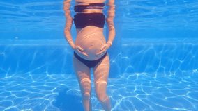 In this slow-motion underwater video, a pregnant woman stands in a swimming pool with her arms resting on her belly. The water gently flows around her. Concept of the benefits of aquatic exercises