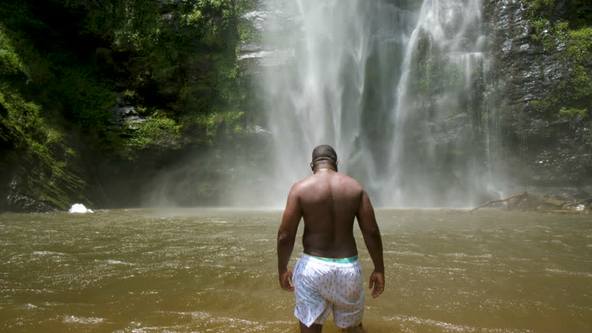 African man walks in a pool of water at the base of a Waterfall