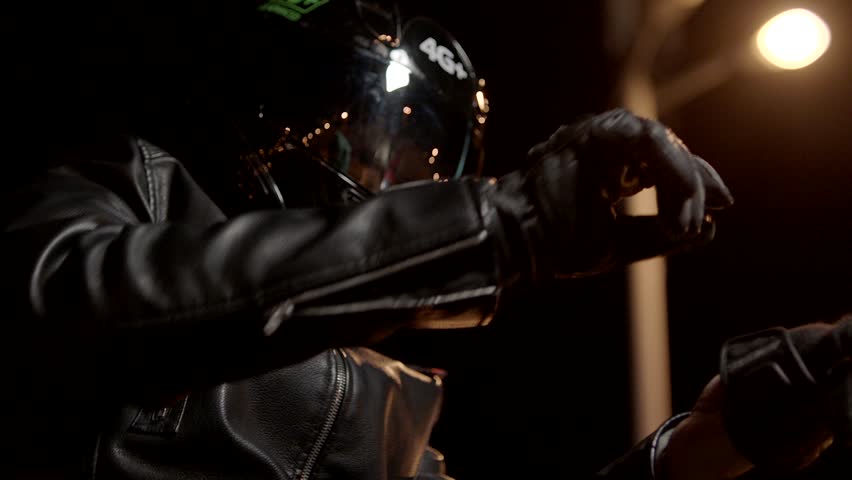 Biker In Leather Jacket, wearing , tying motorcycle black gloves and helmet . Biker Man Taking Off or installing protective clothes . Close up of rider sitting on beautiful motorcycle at night