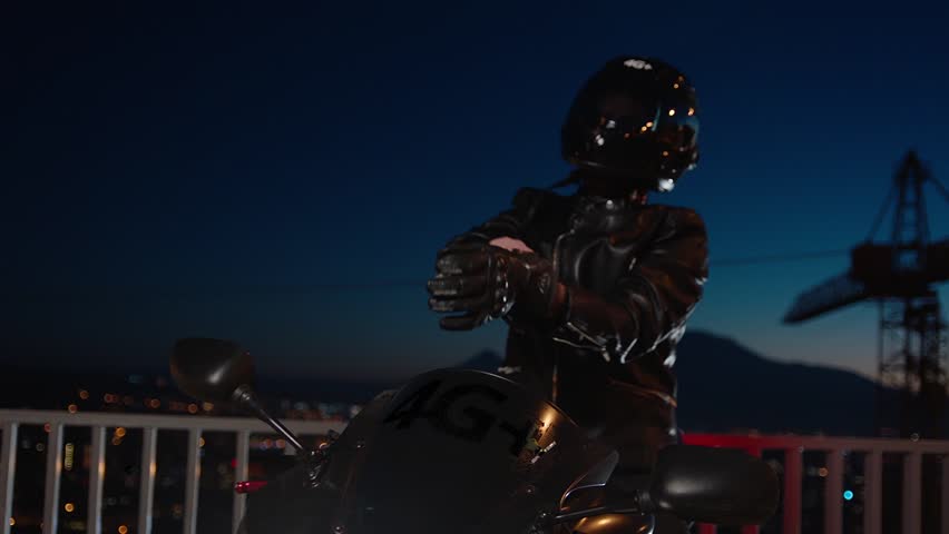 Biker In Leather Jacket, wearing , tying motorcycle black gloves and helmet . Biker Man Taking Off or installing protective clothes . Close up of rider sitting on beautiful motorcycle at night