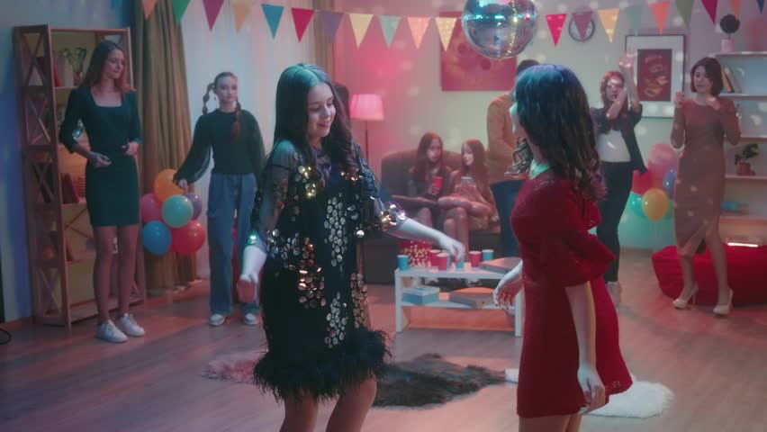 Friends have fun at the party. Teenagers enjoy dancing while girls chat and drink from paper cups while sitting on the couch. Home disco with disco ball and balloons. Slow motion. Royalty-Free Stock Footage #1104062795
