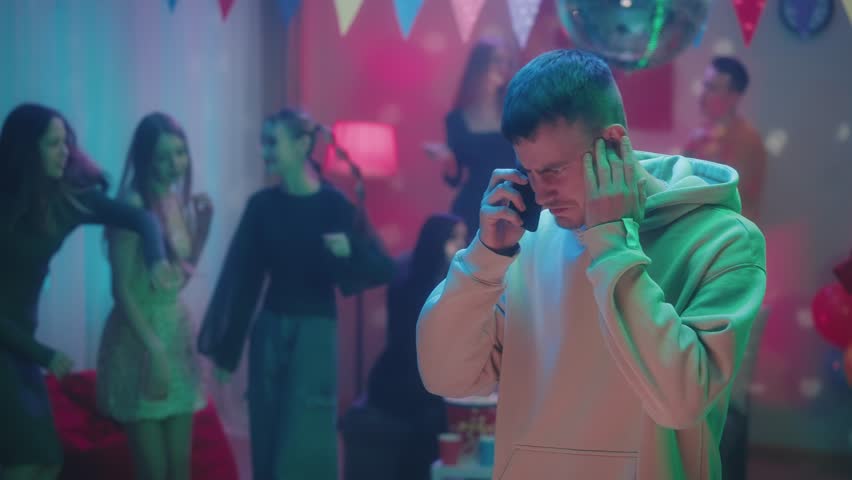 A guy at a party in a decorated room with a disco ball is emotionally talking on the phone while his friends are dancing. He proves something, argues and, upset, hangs up. Slow motion. | Shutterstock HD Video #1104062807