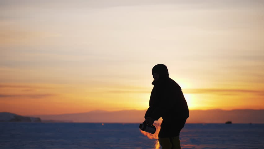 Portrait of person standing on surface of frozen lake at dusk, spilling contents of thermos with beautiful effect of hot water evaporating. Amazing scene demonstration of cool physics effect. Royalty-Free Stock Footage #1104064309