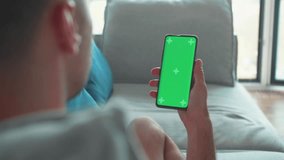 Young Caucasian Man at Home Lying on a Couch using Smartphone with Green Mock-up Screen, Doing Swiping, Scrolling Gestures. Guy Using Mobile Phone, Internet Social Networks Browsing