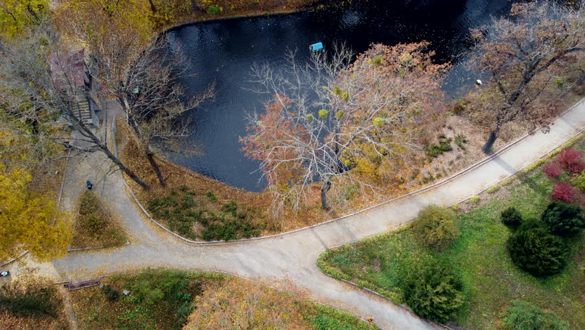 Flying over a blue lake, dirt paths, trees with yellow leaves on the shore of the lake, people in the park on a sunny autumn day. Top view. Beautiful natural background. Aerial drone view.