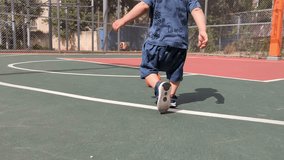 Young infant or toddler running after a ball in an outdoor basketball court near children's playground. Slow motion video of a child playing with a small basketball and kicking it while having fun.