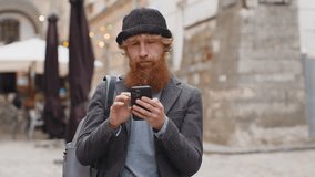 Portrait of young bearded man using smartphone typing text messages looking for a way on map in mobile navigator app outdoors. Redhead guy walking in urban city street background. Town lifestyles