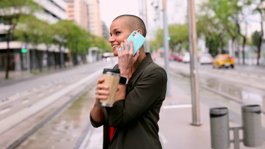 Beautiful mature woman with shaved head casual clothes stands at the tram stop, to travel by public transport, she has a coffee to go and is calling with smart phone