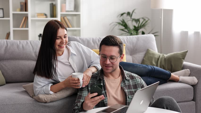 Cheerful young couple relaxing together with laptop and coffee in living room, searching something on phone, having hot drinks, using smartphone and shopping, close up shot. Royalty-Free Stock Footage #1104096117