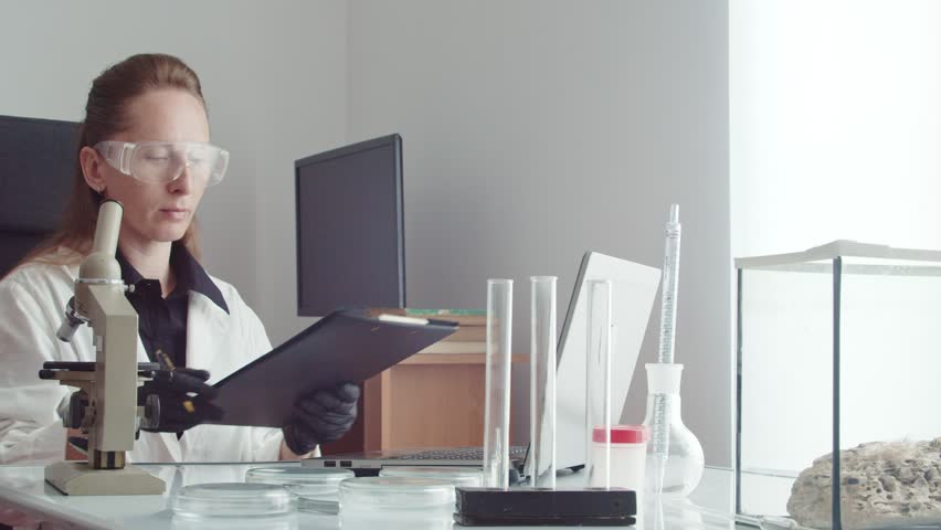Close up portrait of a young adult woman doctor in glasses and a white coat writes in a tablet with a pencil on the background of a laptop and a microscope. The camera moves around the subject