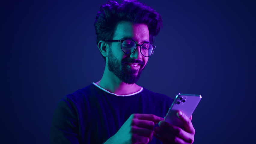 Arabian male using mobile phone chatting browsing smartphone online order shopping gaming with cellphone Indian man scrolling social media internet technology app at neon ultraviolet studio background Royalty-Free Stock Footage #1104100749