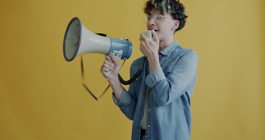 Portrait of young man speaking in megaphone protesting against yellow color background. Social activism and active young people concept.
