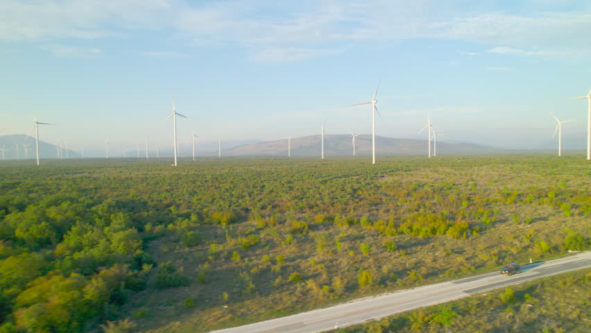 AERIAL: Off-road car passing by working wind turbines in beautiful sunset light. Picturesque view of wind farm with scattered rotating windmills and roadway leading through Mediterranean landscape. Royalty-Free Stock Footage #1104102961