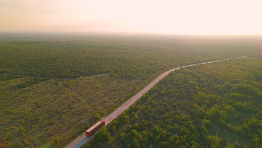 AERIAL: Red container truck driving along Adriatic landscape in golden sunlight. Big delivery truck transporting goods through beautiful countryside with lush green shrubbery in early morning light. Royalty-Free Stock Footage #1104102965