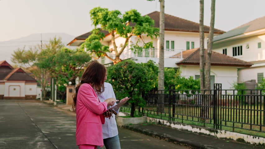 A real estate agent and her client are having a light-hearted conversation and joking around while standing in front of houses in a high-end neighborhood