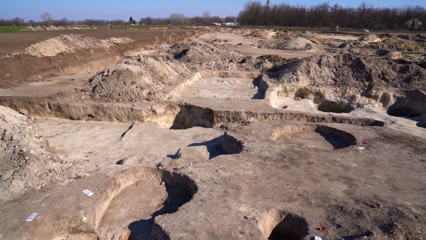 High angle establishing view of archaeology dig site marked with pits, dirt mounds.