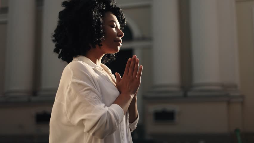 Portrait of hopeful young African American woman clasping hands in prayer asking for blessing and help while the rays of rising sun fall on her face near church outdoors Religion and faith concept Royalty-Free Stock Footage #1104119299
