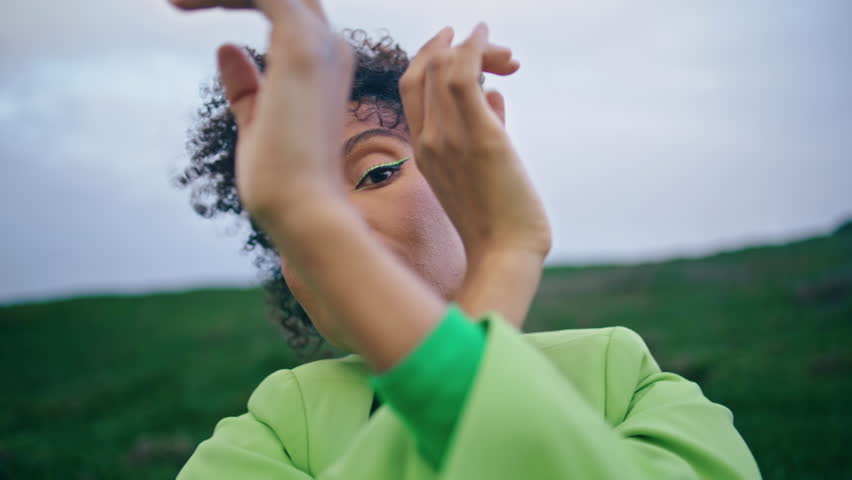 Closeup african american young woman dancing sensually in front cloudy gray sky. Portrait inspired emotional girl moving hands smoothly standing on green field. Attractive lady dancer looking camera.