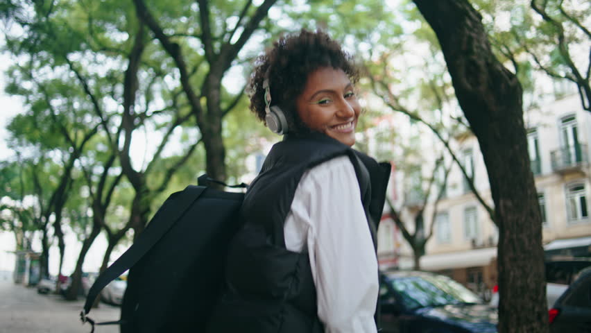 Joyful funky woman walking dancing along listening music in wireless headphones outdoors. Happy african american girl have fun enjoying sound in modern earphones. Smiling lady tourist strolling street