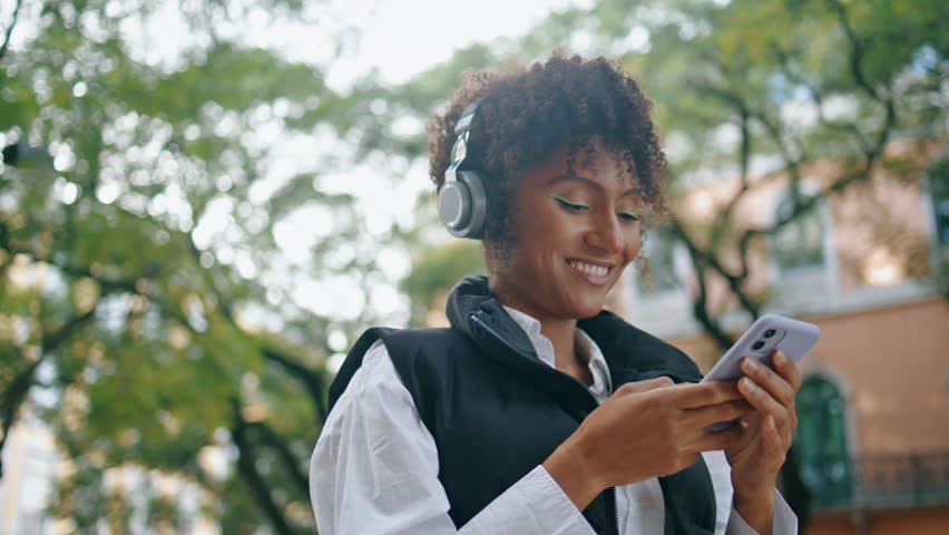 Cheerful african american lady listening music by wireless headphones holding mobile phone close up. Attractive happy woman using telephone app standing city street. Positive girl enjoy favorite song.
