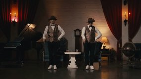 Two stylish professional male dancers in classy dandy hat performing jazz funk dance synchronic moves in vintage production studio retro environment with theatrical red velvet curtains and candlelight