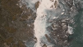 Drone video moving from top down, white waves flowing over large rocks with blue water.