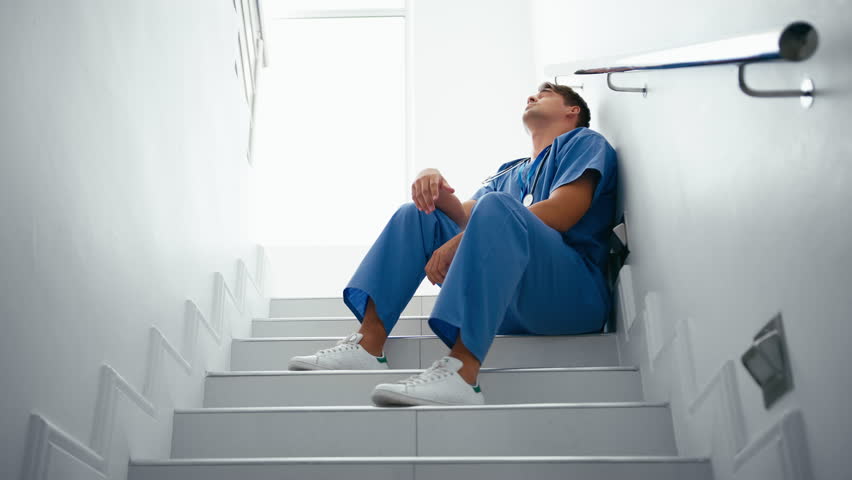Stressed and tired male doctor or nurse wearing scrubs sitting on hospital stairs - shot in slow motion Royalty-Free Stock Footage #1104136457
