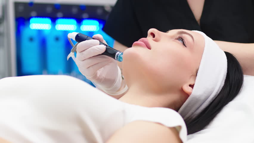 View of doctor cosmetologist doing anti aging procedure in cosmetology office. Satisfied woman in disposable hat lying on couch and relaxing. Working with Apparatus. Royalty-Free Stock Footage #1104138569