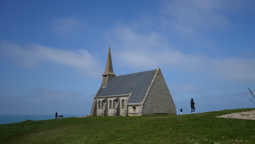 Unrecognizable people walking around small old chapel in Etretat found on top of cliff fronting big ocean, on background of blue cloudy sky. Concept of vacations in France. Shooting in slow motion.