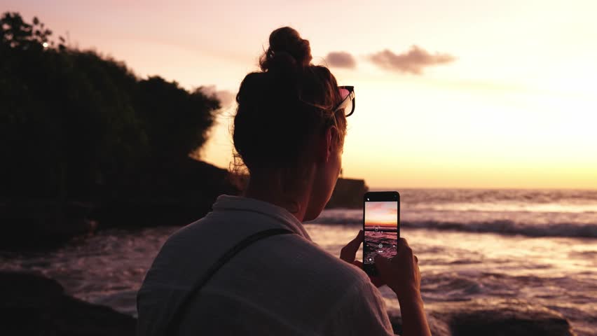 Woman Sitting Outdoors at Sea Beach Holding Mobile Phone to Make Picture. Hipster Girl at Summertime Break on Coastline Side Taking Footage for Social Media. Female in Ocean Wind of Seashore Nature Royalty-Free Stock Footage #1104148619