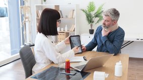 Back view of female doctor holding digital tablet while sitting in front of smiling caucasian man in cervical collar. Health professional consulting mature patient about neck injury using CT scan. - Powered by Shutterstock - Get 15% off with code: PIKWIZARD15