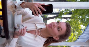 Online video call with relatives or friends during coffee break. Young woman hold her smartphone while sitting at sidewalk cafe and talking. Slow motion vertical 4k footage.