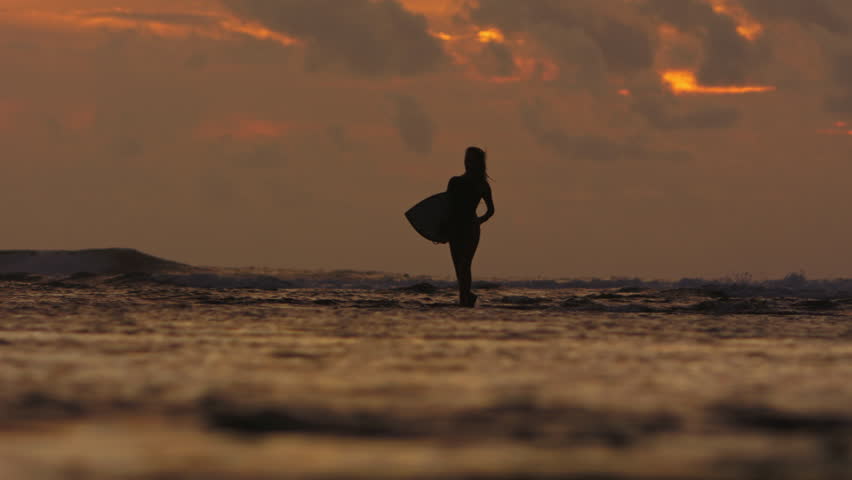 Silhouette of sexy slender female woman surfer carrying longboard and walking on warm ocean water at orange summer sunset. Sexy seductive silhouette of female surfer with longboard in her hand.