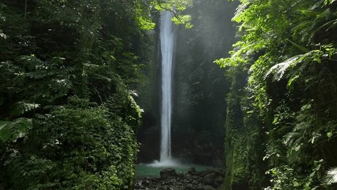 Drone shoot. Waterfall hidden in tropical rainforest jungle,  falling water hitting water surface, some huge rocks seeable in through splashes. Lush green leaves is moving from the wind. Philippines Video Stok