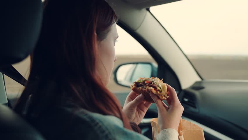 Girl passenger eats burger in car. Delicious food bun with cutlet. Young hungry woman eats roll with meatball while sitting in car. Teenager travels, having snack on road without leaving car.Tourist Royalty-Free Stock Footage #1104170617